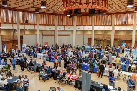 high angle, looking down at a large open auditorium with dozens of people talking around different job fair vendor tables and displays